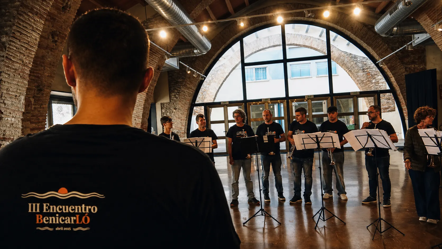 Participante de espaldas con la camiseta del III Encuentro Benicarló, frente al grupo de trompetas durante un ensayo